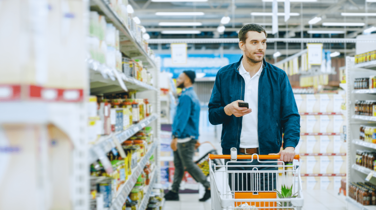 a caucasian man grocery shopping. He's holding his phone in his right hand and pushing a groery cart down an aisle with his left.