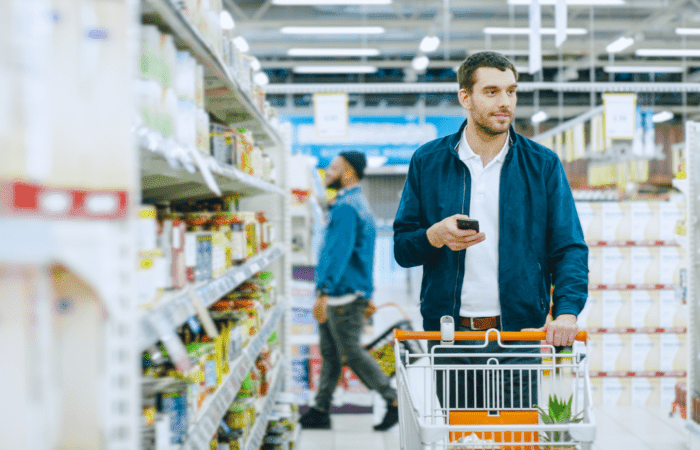 a caucasian man grocery shopping. He's holding his phone in his right hand and pushing a groery cart down an aisle with his left.