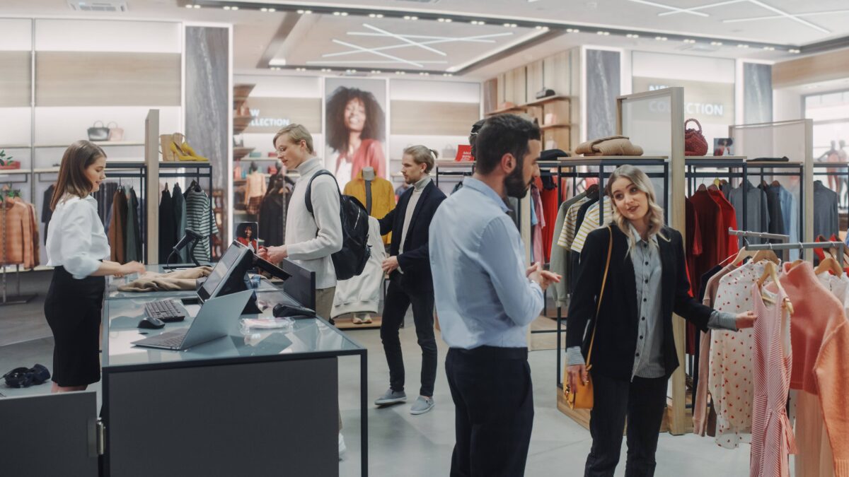 a retail boutique, with a line for the registers and an employee helping, and a woman getting help from an employee off to the side