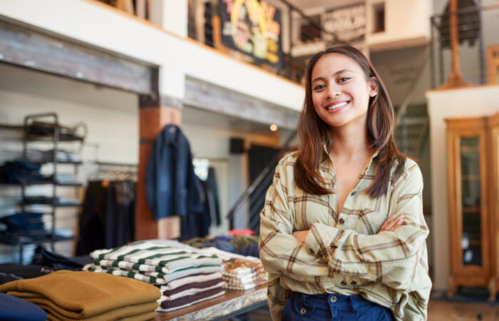 Portrait Of Smiling Female Owner Of Fashion Store Standing In Front Of Clothing Display