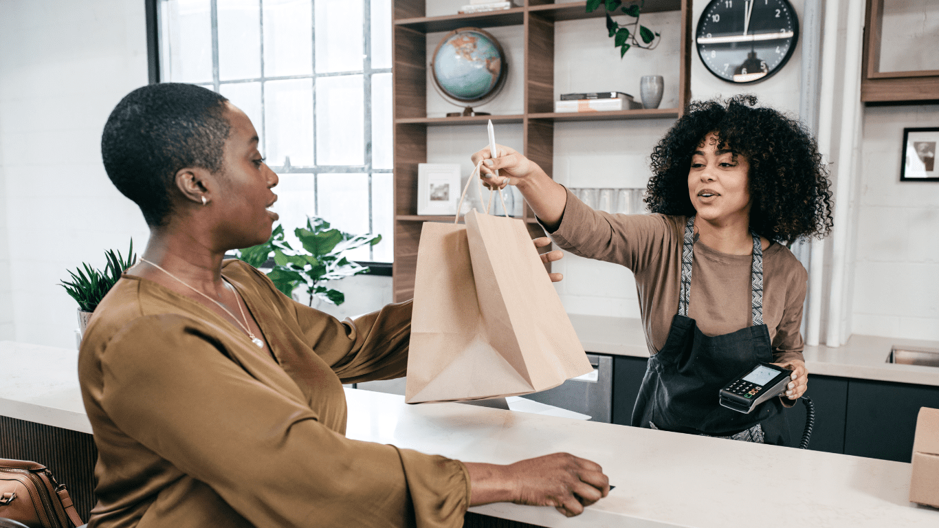 an african american employee smiling and handing a paper shopping bag over the counter to a customer