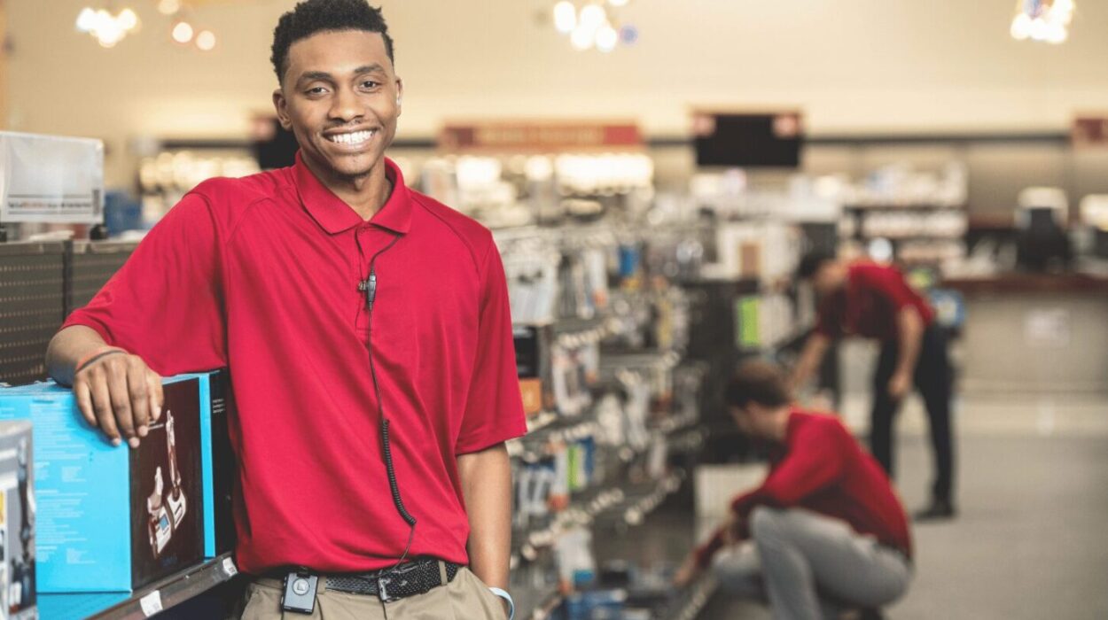 retail employee smiling, wearing red polo shirt with arm resting on shelf. wearing a theatro mobile communication platform device