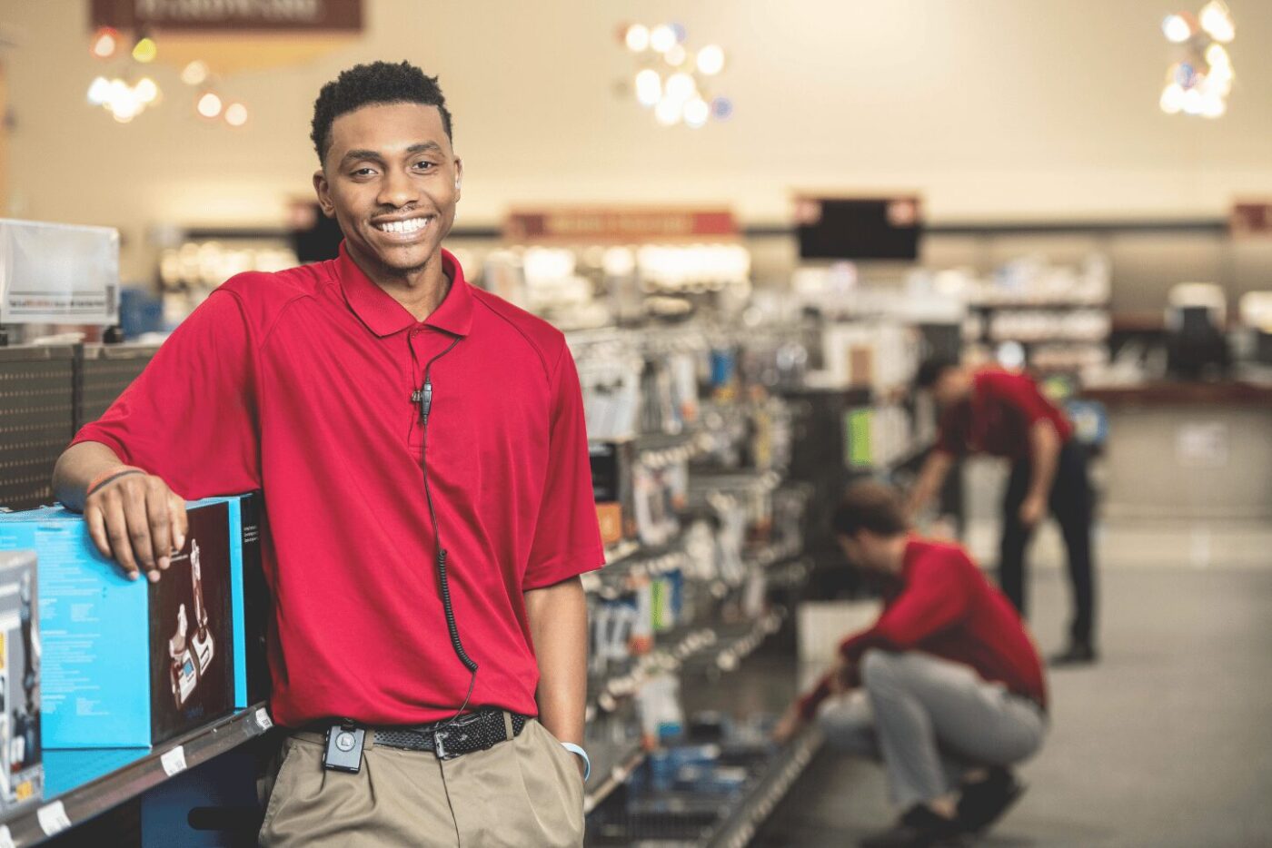 retail employee smiling, wearing red polo shirt with arm resting on shelf. wearing a theatro mobile communication platform device